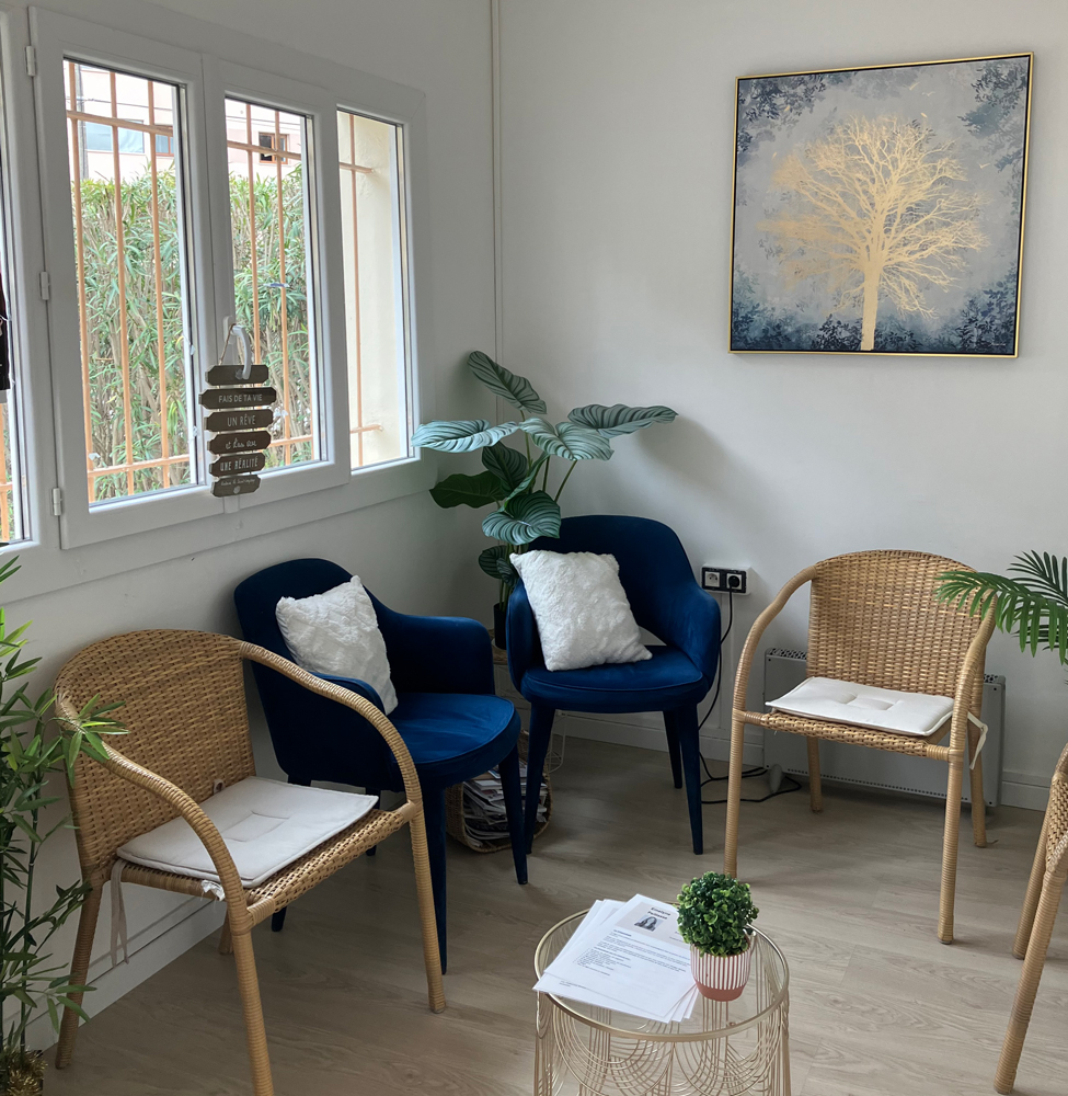 Photo montrant la salle d'attente du cabinet thérapeutique de Sophie Ianni à Mandelieu, avec une grande fenêtre, un tableau avec un arbre doré sur fond bleu, des chaises en rotin, des fauteuils en velours bleu marine et quelques plantes