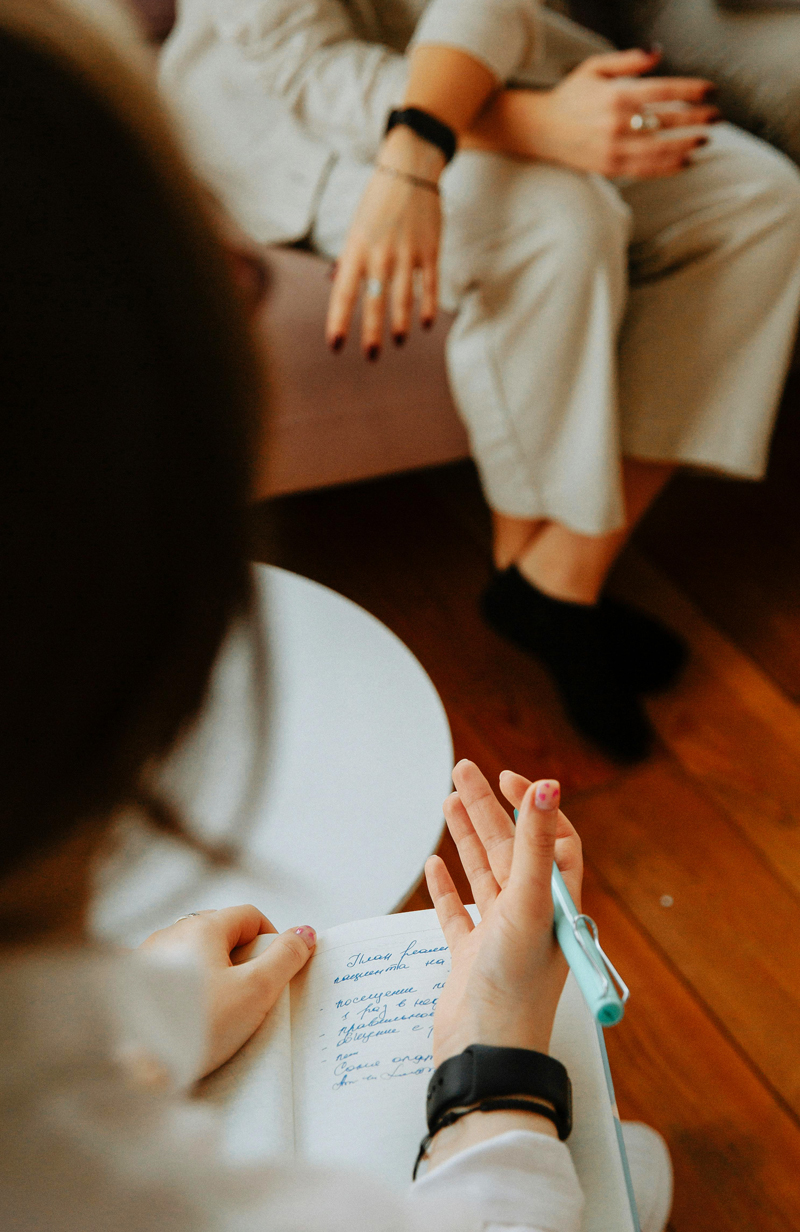 Photo montrant deux femmes assises l'une en face de l'autre : celle au premier plan est une thérapeute, de dos, qui prend des notes, tandis qu'en face se trouve sa patiente, assise sur un canapé, les pieds croisés, en chaussettes